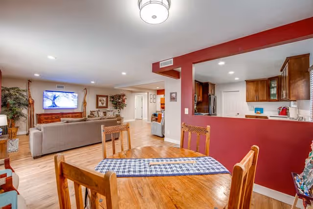 Open-plan common area with a wooden dining table in the foreground, a living room with sofas and a wall-mounted TV, and a kitchen behind a red half-wall.