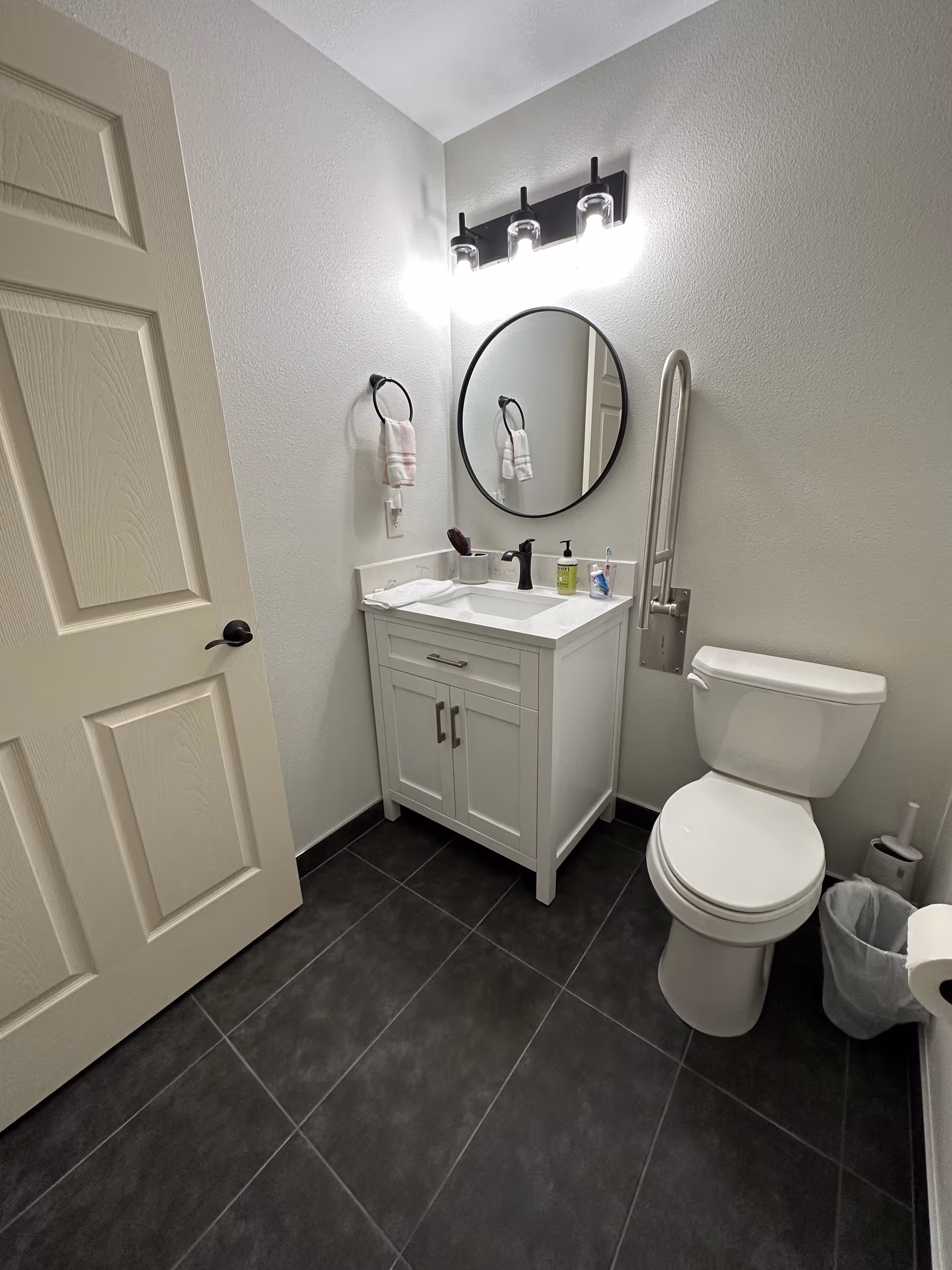 A clean and modern bathroom with a white vanity cabinet, a round mirror above the sink, a black faucet, a towel ring with a hand towel, a toilet with a grab bar mounted on the wall, a trash can, and a toilet paper holder. The floor is covered with dark tiles and the walls are painted light gray.