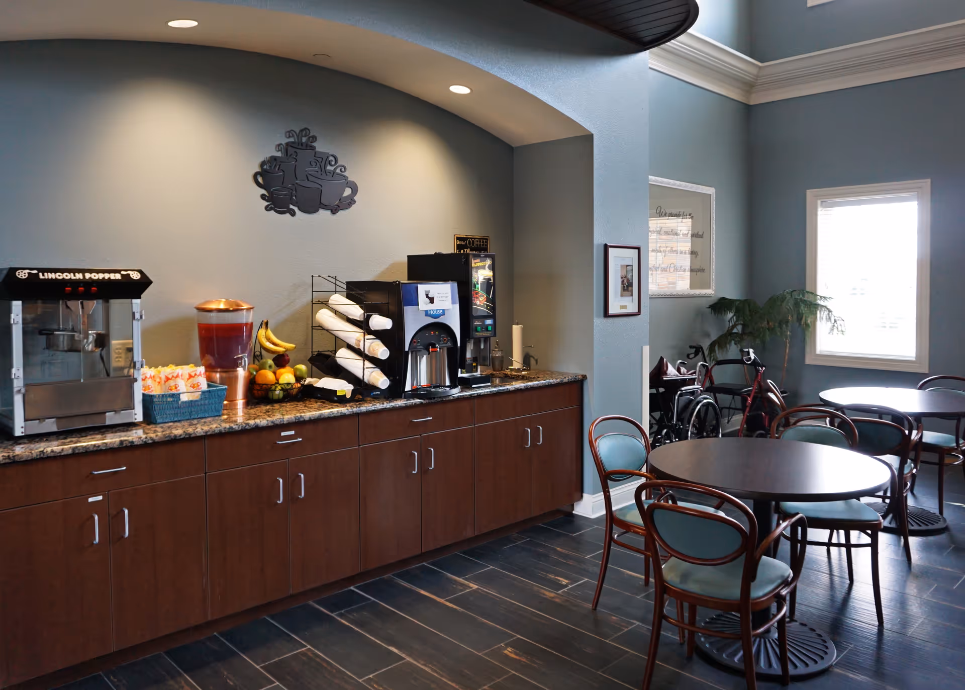 A cozy dining area in Snyder Village Assisted Living featuring round tables with chairs, a beverage station with a popcorn machine, juice dispenser, coffee machine, and fruit bowl, set against a light blue wall with decorative wall art and a window letting in natural light.