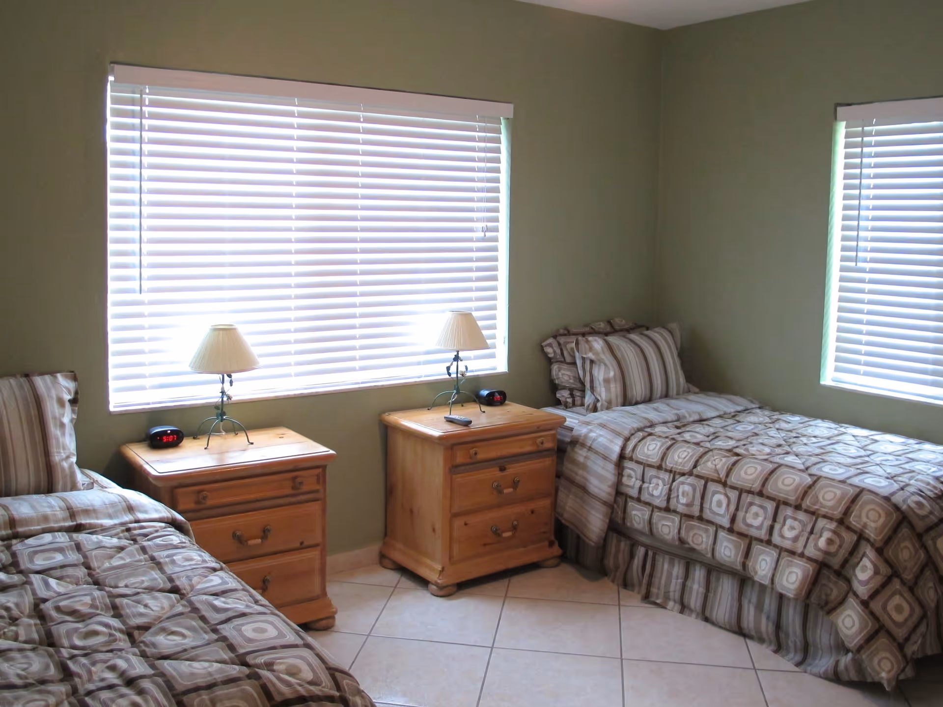 Two twin beds with patterned bedspreads, wooden nightstands and lamps beneath large window blinds in a small bedroom.