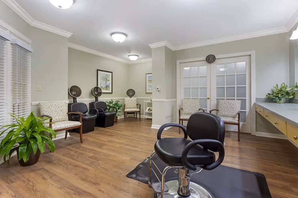 Interior view of a hair salon area in a senior living facility with wooden flooring, multiple salon chairs including black styling chairs and beige patterned waiting chairs, plants, framed artwork on the walls, and a counter with drawers on the right side.