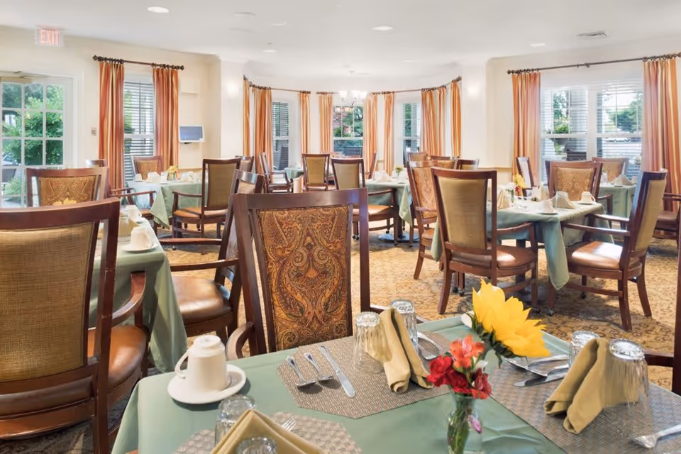 A bright and spacious dining room with multiple tables covered in green tablecloths, each set with cups, utensils, napkins, and small flower arrangements. The room features large windows with orange curtains, wooden chairs with patterned upholstery, and a carpeted floor.