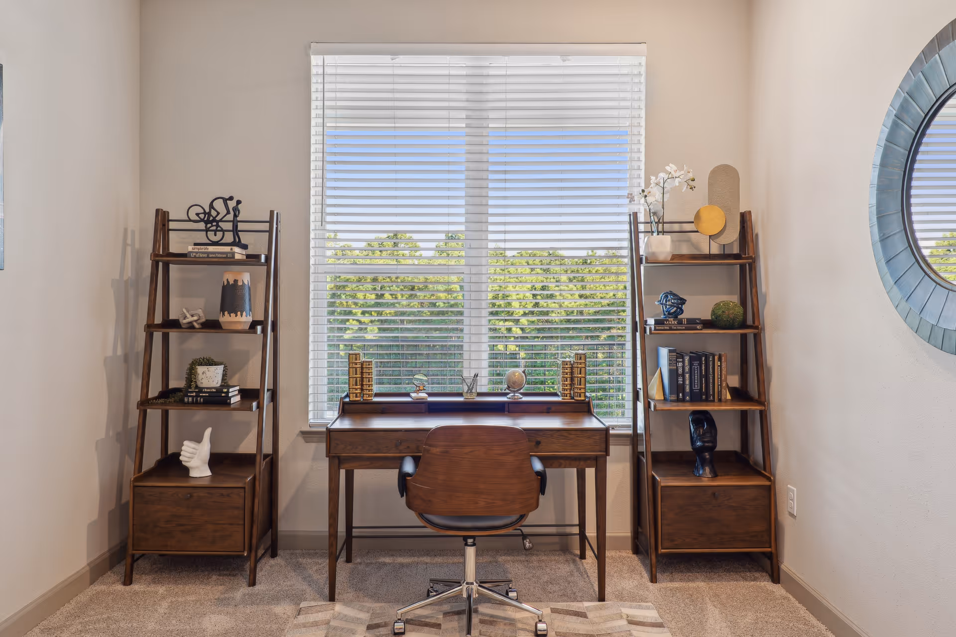 A home office setup with a wooden desk and a swivel chair positioned in front of a window with white blinds. On either side of the desk are two wooden ladder-style shelves holding decorative items and books. A round mirror is mounted on the wall to the right.