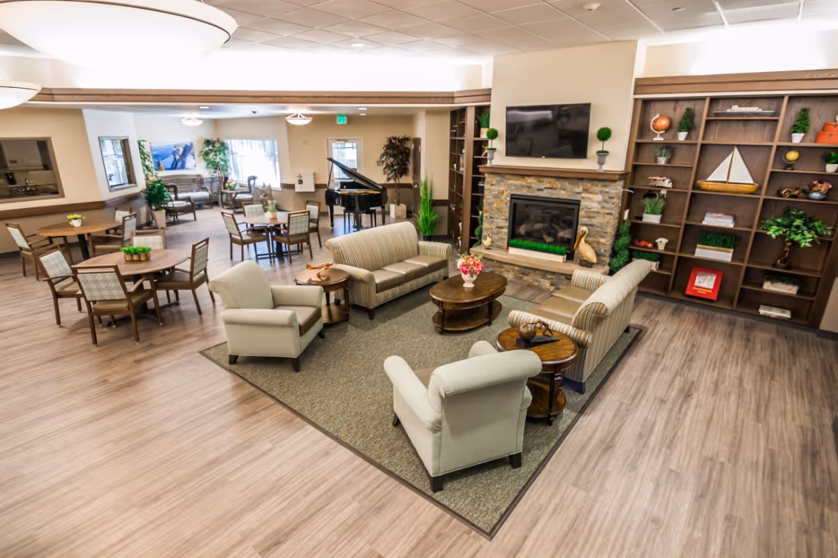 A spacious and well-lit common area in a senior living facility featuring a seating arrangement with two sofas and two armchairs around a coffee table on a rug. Behind the seating area is a stone fireplace with a mounted flat-screen TV above it and built-in wooden shelves decorated with plants, books, and ornaments. To the left, there are several round tables with chairs, a grand piano, and large windows letting in natural light.