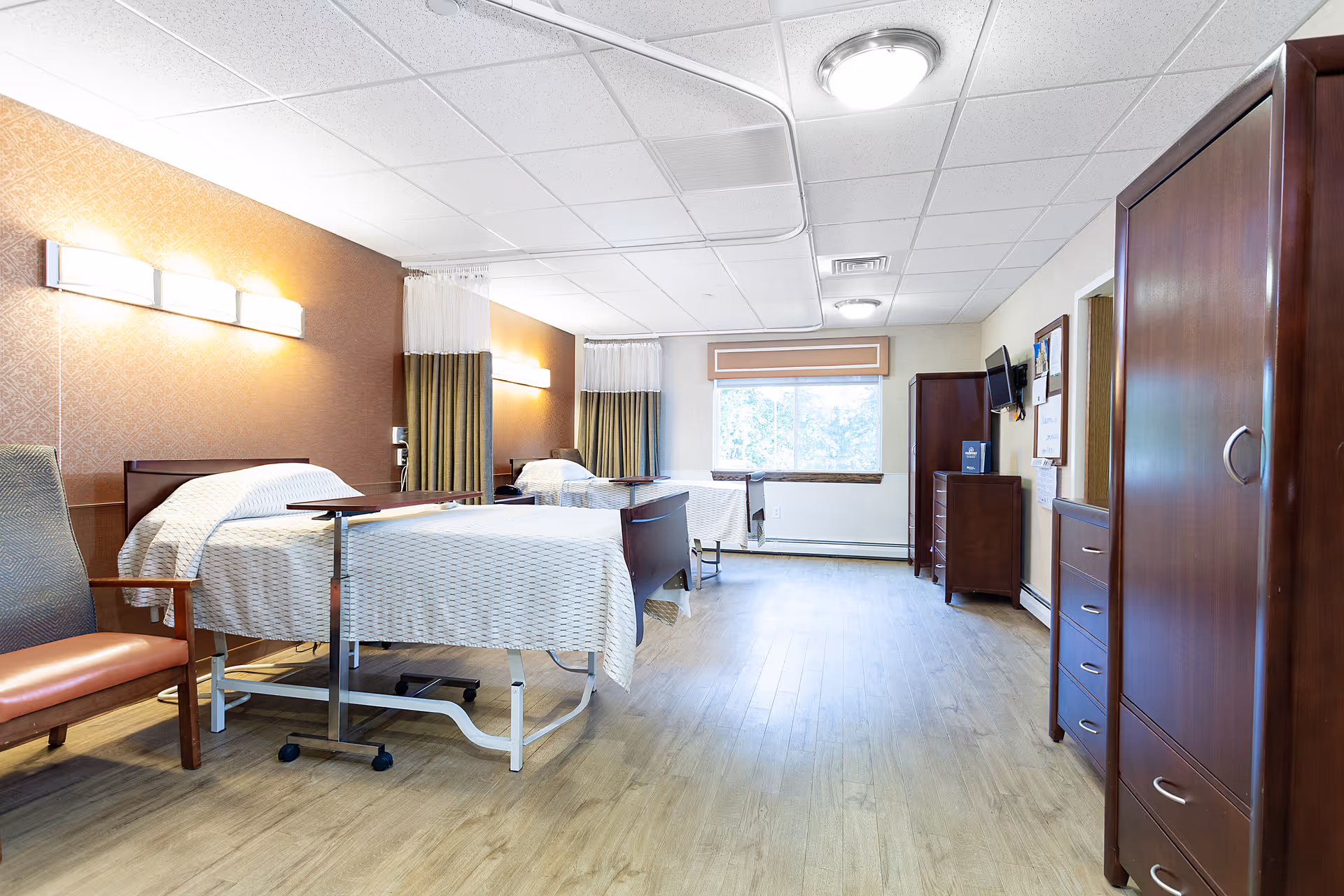 A bright and clean patient room in a healthcare facility with two hospital beds separated by privacy curtains. The room has wooden flooring, a large window with a beige valance, wooden furniture including a wardrobe, dresser, and chair, and wall-mounted lights above each bed.