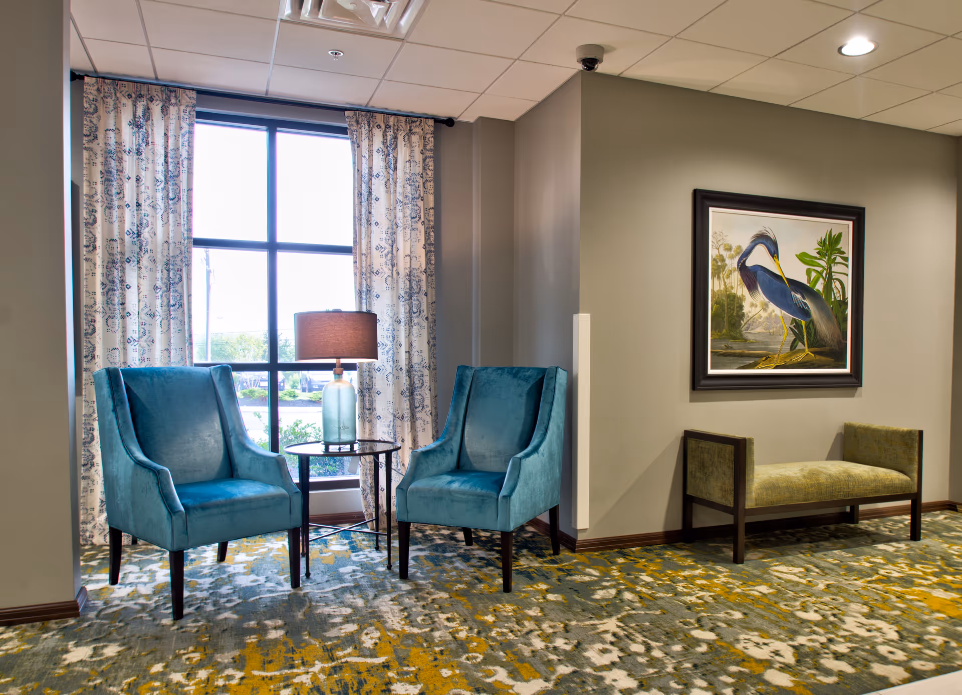 A cozy seating area with two teal upholstered armchairs and a small round glass table with a lamp between them, positioned in front of a large window with patterned curtains. To the right, there is a green cushioned bench beneath a framed painting of a heron bird. The floor is covered with a patterned carpet in shades of green, yellow, and white.