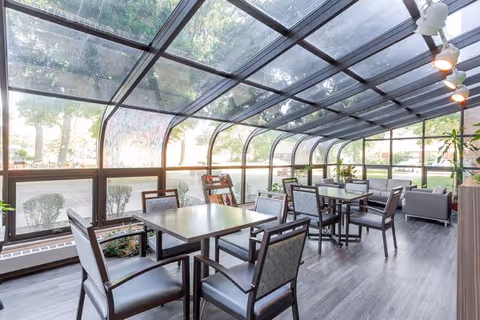 Sunlit enclosed dining area with dark wood tables, chairs, and potted plants.