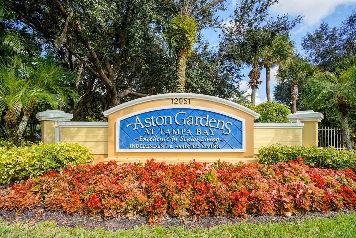 Entrance sign for Aston Gardens at Tampa Bay surrounded by lush greenery and colorful flowers with trees in the background under a partly cloudy sky.