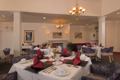Elegant dining room with multiple round tables dressed in white linens, place settings with folded red napkins, floral centerpieces, chandeliers, and upholstered chairs.