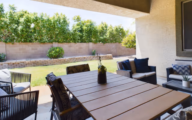Covered outdoor patio area with a large wooden table surrounded by cushioned chairs. The patio overlooks a green lawn with a stone retaining wall and a row of trees along a brick fence under a clear blue sky.