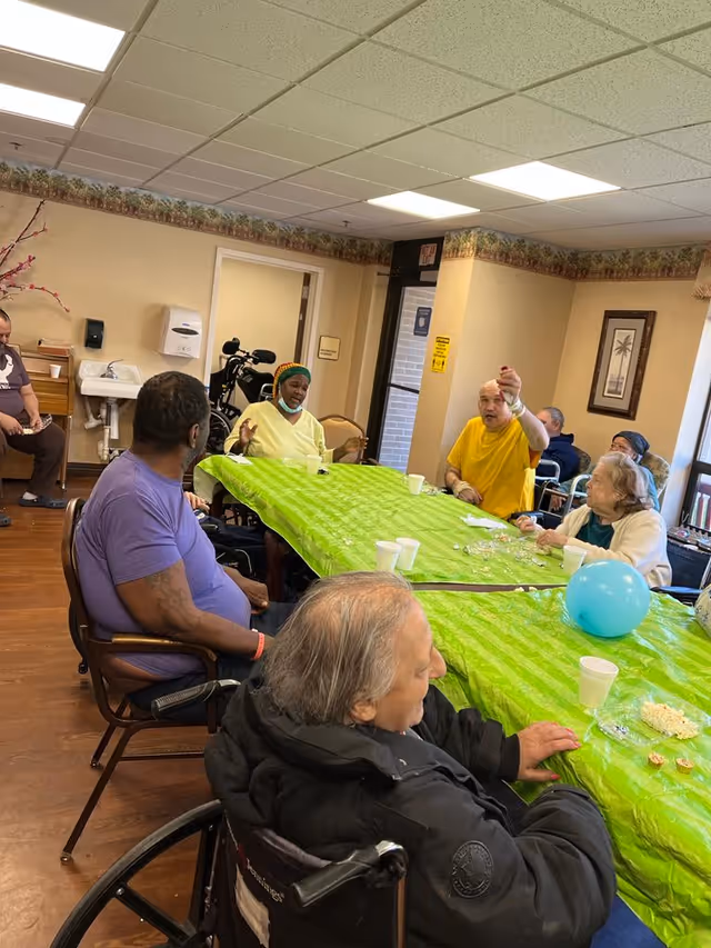 A group of elderly people sitting around a long table covered with a bright green tablecloth in a common room. Some individuals are in wheelchairs, and there are cups, plates with snacks, and a blue balloon on the table. The room has beige walls, wood flooring, and fluorescent ceiling lights. A sink and hand sanitizer dispenser are visible on the wall, along with framed artwork and a door leading outside.