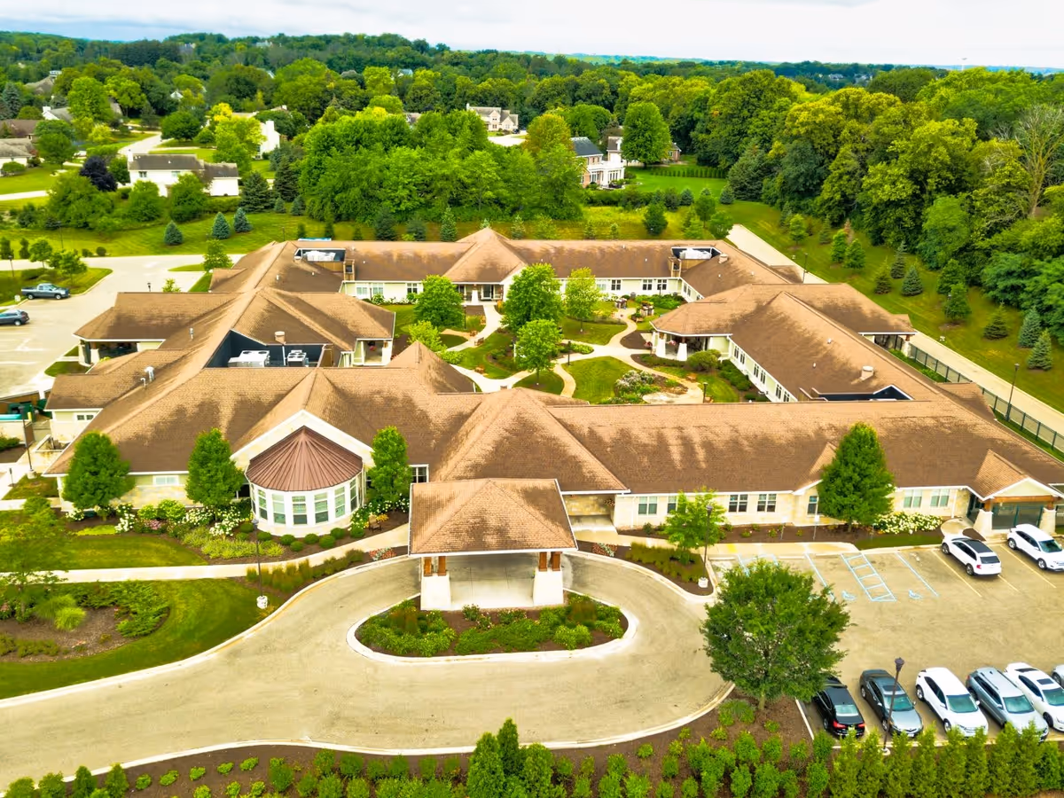 Aerial view of The Auberge at Brookfield senior living facility showing a large, single-story building with a brown roof arranged in a U-shape around a landscaped courtyard with trees and pathways. The building is surrounded by green lawns, trees, and a parking lot with several cars parked.