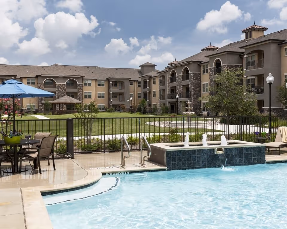Outdoor swimming pool area with clear blue water, a small water feature with fountains, poolside chairs, and a table with an umbrella. In the background, there is a large multi-story residential building with balconies, green lawn, trees, and a gazebo under a partly cloudy sky.