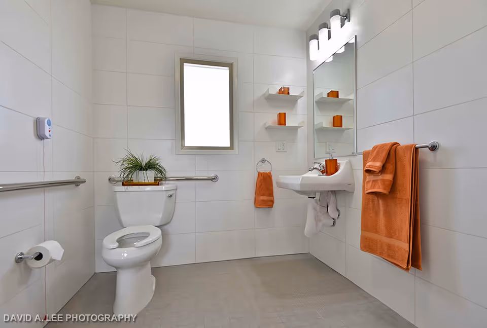 A clean, accessible bathroom with white tiled walls and floor. It features a white toilet with a grab bar on the wall behind it, a small plant on top of the toilet tank, a wall-mounted sink with orange towels hanging on a towel ring and a towel bar. Above the sink is a mirror with a light fixture and two small shelves holding orange decorative items. There is a window on the back wall allowing natural light.