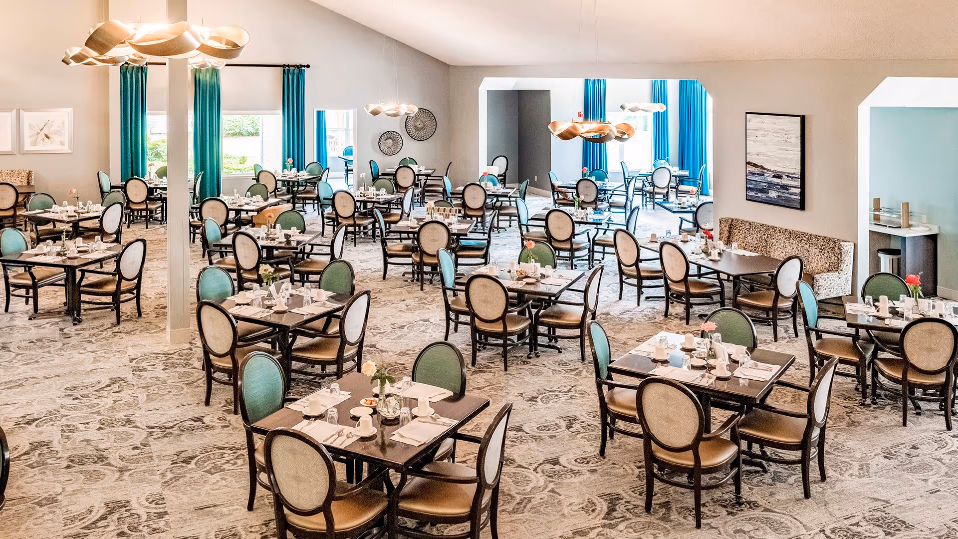 A spacious dining room with multiple tables set for meals, each table surrounded by chairs with light-colored upholstery. The room features large windows with teal curtains, modern pendant lighting, and light-colored walls with decorative artwork. The carpet has a subtle patterned design.