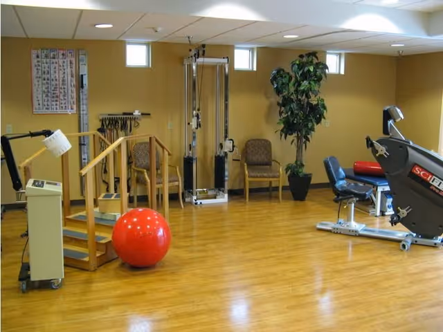 A rehabilitation or physical therapy room with exercise equipment including a red exercise ball, parallel bars, a seated exercise machine, chairs, a potted plant, and a chart on the wall. The room has wooden flooring and beige walls with small windows near the ceiling.