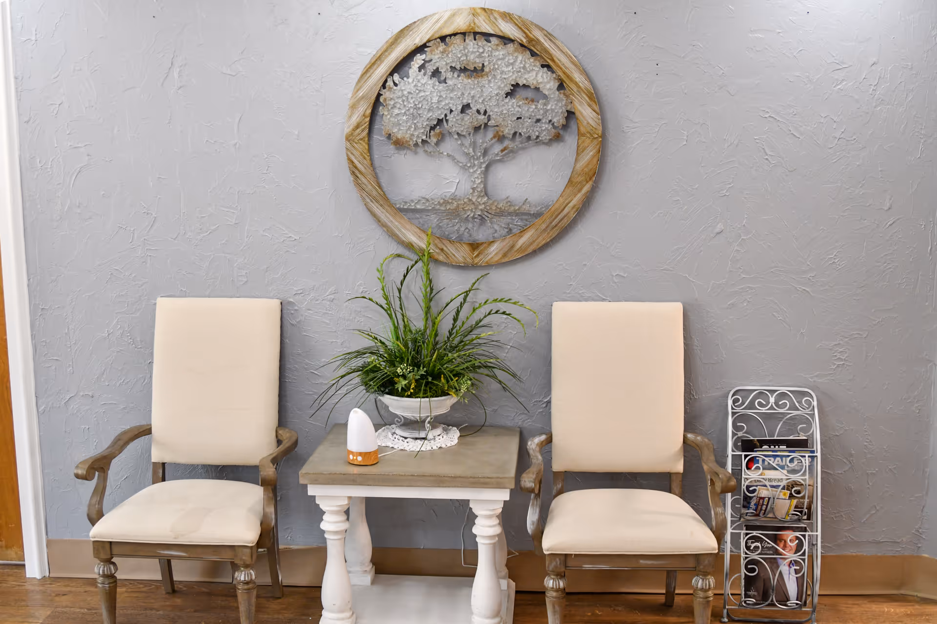 Two beige upholstered armchairs with wooden frames are placed on either side of a small table with a potted plant and a small device on it. Behind the table on the gray textured wall is a round wooden wall decoration featuring a tree design. To the right of the chairs is a metal magazine rack holding several magazines.