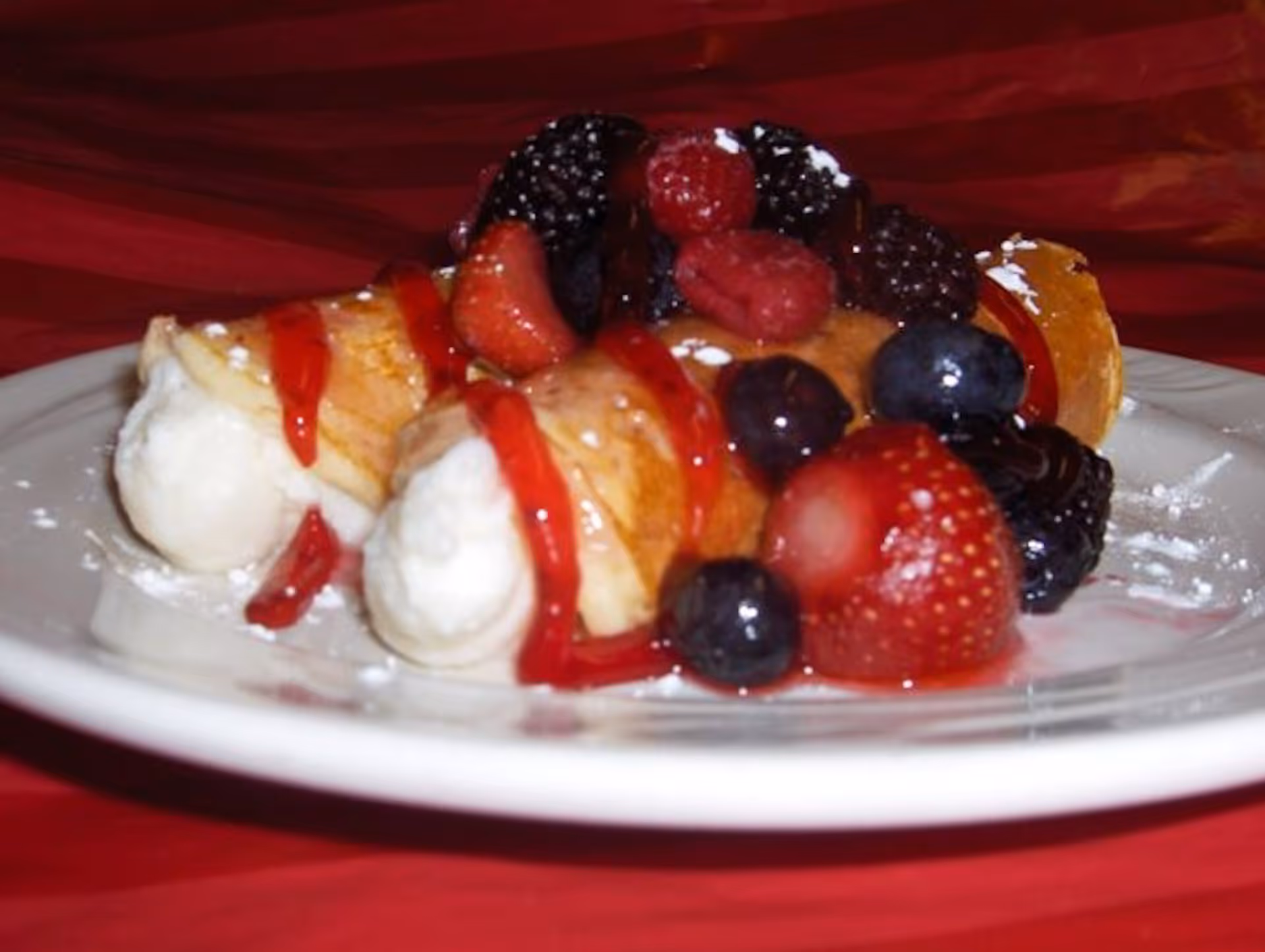 A white plate with two cannoli pastries filled with cream, drizzled with red sauce, and topped with assorted fresh berries including strawberries, blueberries, raspberries, and blackberries, on a red tablecloth background.