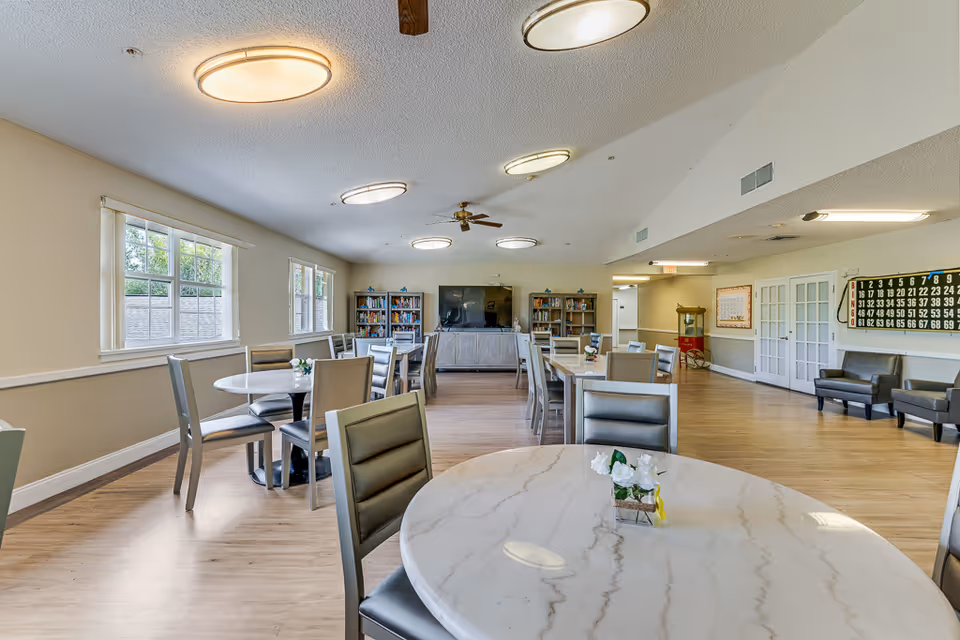 A spacious senior living facility common area with several round and rectangular tables surrounded by chairs. The room has large windows letting in natural light, bookshelves with books, a large flat-screen TV, ceiling fans, and modern ceiling lights. There are also a few armchairs along the wall and a bingo board on the far wall.