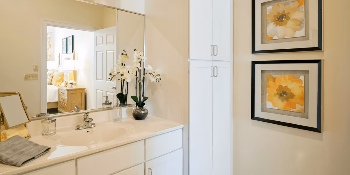 A bathroom vanity with a white countertop, sink, and silver faucet. On the countertop are a small mirror, a glass container, a soap dispenser, a vase with white orchids, and folded towels. A large mirror above the sink reflects a bedroom with a bed, nightstand, and lamp. On the wall next to the vanity are two framed floral artworks with yellow flowers. White cabinets are visible beside the vanity.