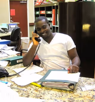 A man wearing a white shirt is sitting at a desk in an office environment, talking on a corded telephone. The desk is cluttered with papers, a binder, and office supplies. Behind him are shelves filled with binders and office materials.