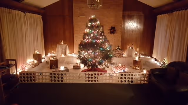A cozy room decorated for Christmas with a lit Christmas tree in the center surrounded by small lit decorations and wrapped presents. The room has wood-paneled walls, cream-colored curtains, and a dark leather armchair on the right side.