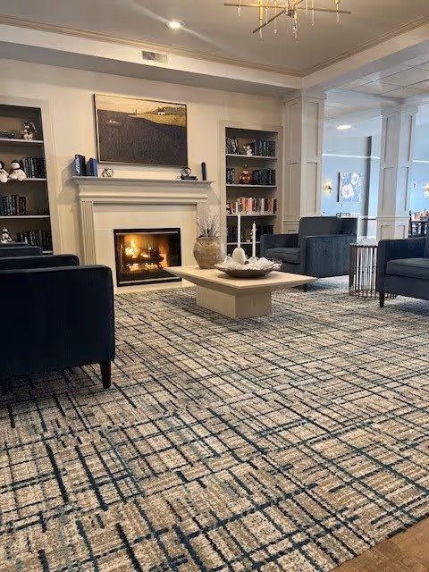 Senior living common room with armchairs around a coffee table in front of a lit fireplace and built-in bookshelves.