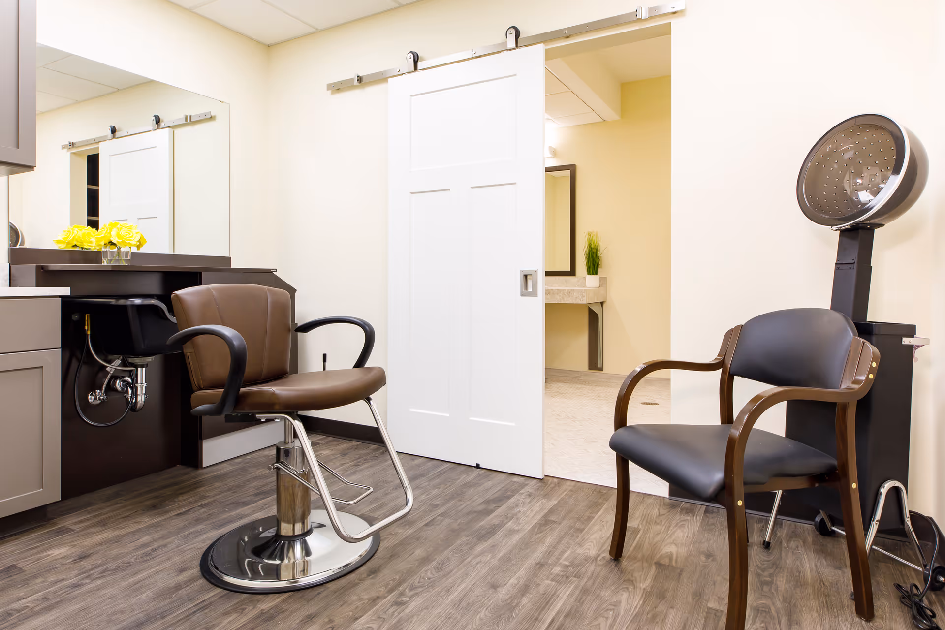 An interior salon area with a styling chair, hooded hair dryer, sink, mirror and a sliding door.