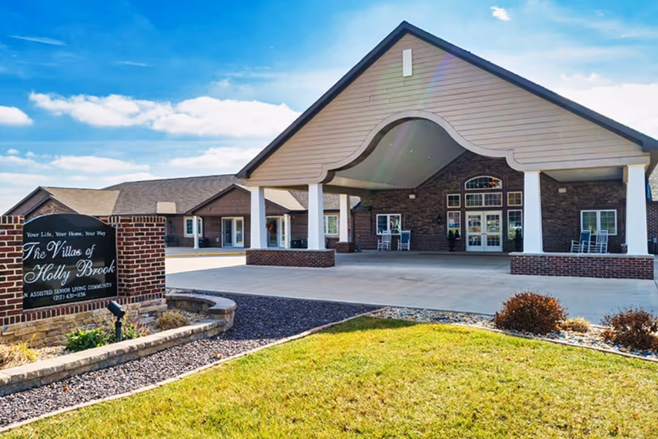 Exterior front view of Monticello Villas of Holly Brook, an assisted senior living community, showing a large covered entrance with white pillars, brick walls, and a sign with the community name on a landscaped lawn under a blue sky with some clouds.