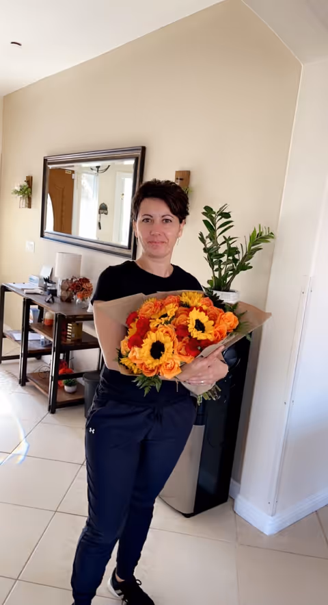 A woman standing indoors holding a large bouquet of orange and yellow flowers. She is dressed in a black top and dark pants. Behind her is a beige wall with a large mirror, a small table with various items, and a green potted plant.