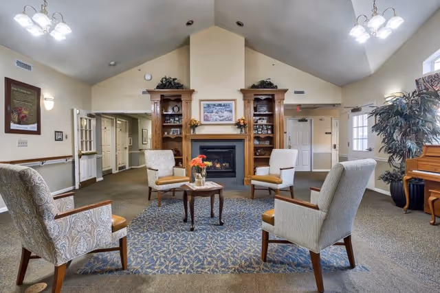 Common room with four upholstered armchairs around a central table on a rug, facing a fireplace flanked by built-in wooden shelves.