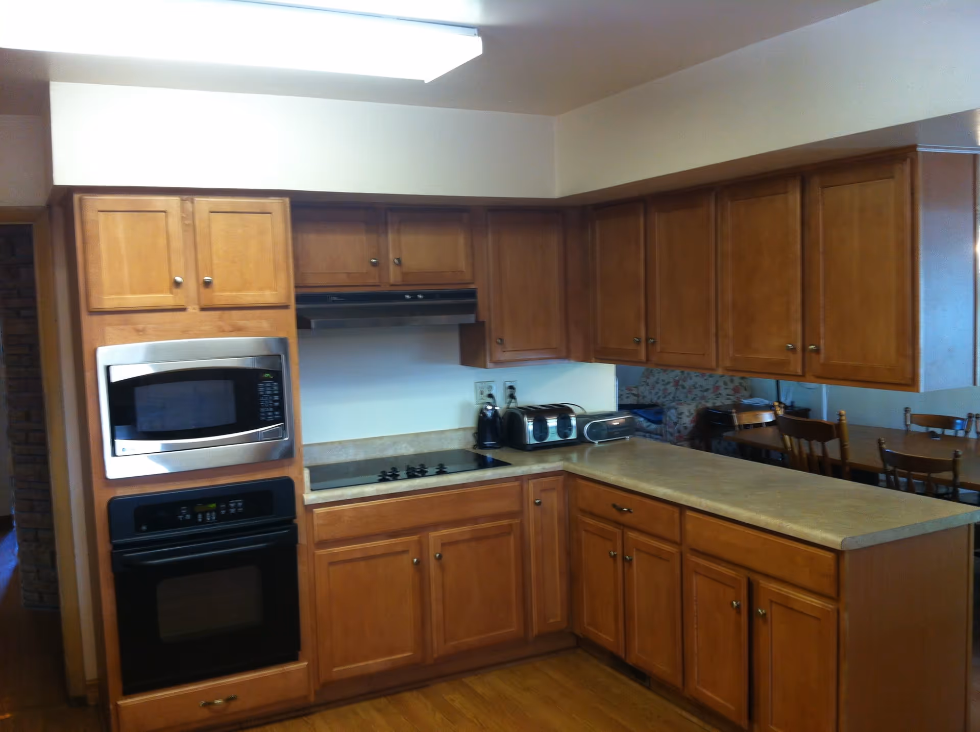 A kitchen area with wooden cabinets, a built-in microwave and oven, an electric stovetop, and a countertop with a toaster, coffee maker, and radio. In the background, there is a dining area with wooden chairs and a floral-patterned couch.