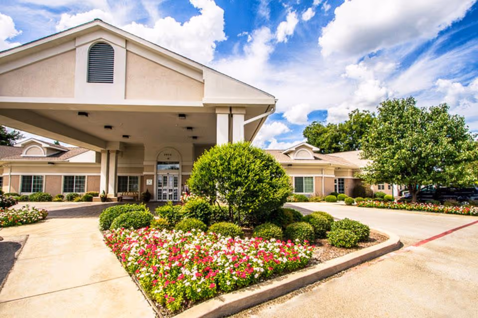 Front exterior view of Life Care Center of Haltom showing a covered entrance with columns, landscaped flower beds with pink and white flowers, green bushes, and trees under a partly cloudy blue sky.