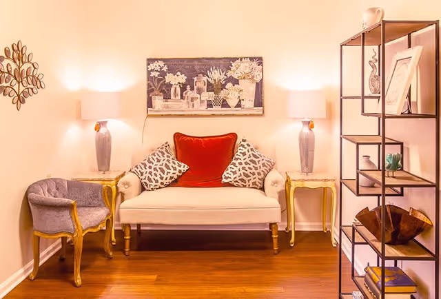 A cozy living room area with a beige loveseat featuring a red cushion and two patterned pillows, flanked by two side tables each with a lamp. To the left is a purple upholstered armchair with wooden legs. On the right side, there is a black metal and wood shelving unit holding decorative items and books. The walls are light-colored with a floral painting above the loveseat and a decorative metal wall art piece on the left.