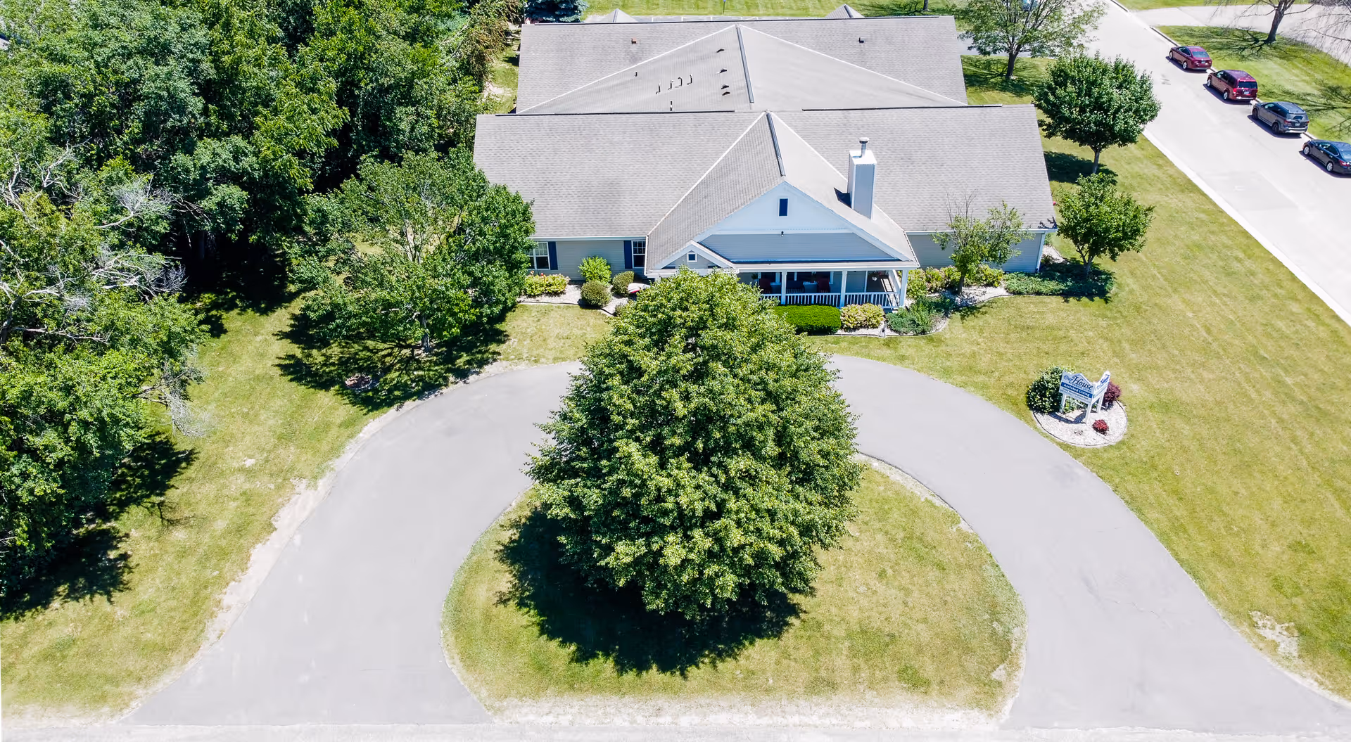Aerial view of a single-story assisted living building with a circular driveway, large tree in front, and surrounding lawns and trees.