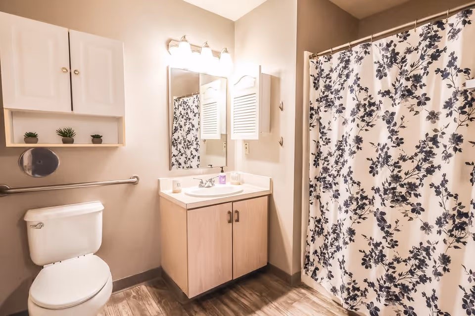 A clean bathroom featuring a white toilet, a wooden vanity with a sink and mirror above it, a wall-mounted cabinet with small potted plants, and a shower with a floral-patterned curtain.