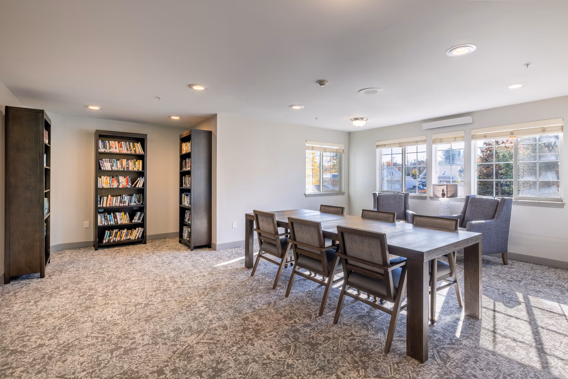 A bright and spacious room with large windows letting in natural light. The room features a long wooden table with six chairs, two upholstered armchairs near the windows, and three tall bookshelves filled with books against the wall. The carpet has a subtle patterned design, and the walls are painted white.