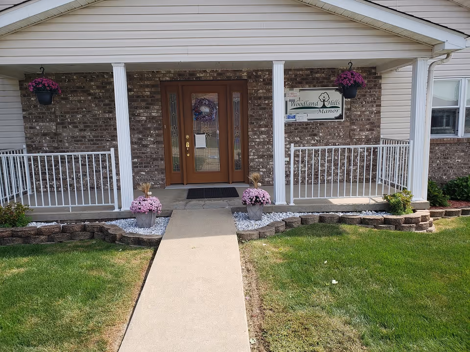 Front entrance of Woodland Hills Assisted Living featuring a covered porch with white railings, two white columns, a brown double door with decorative glass panels, hanging flower pots with purple flowers, and flower pots with pink flowers on either side of the concrete walkway leading to the door.