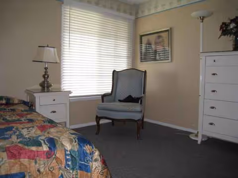 A bedroom in a senior living facility with bed, dresser, chair, and window blinds.