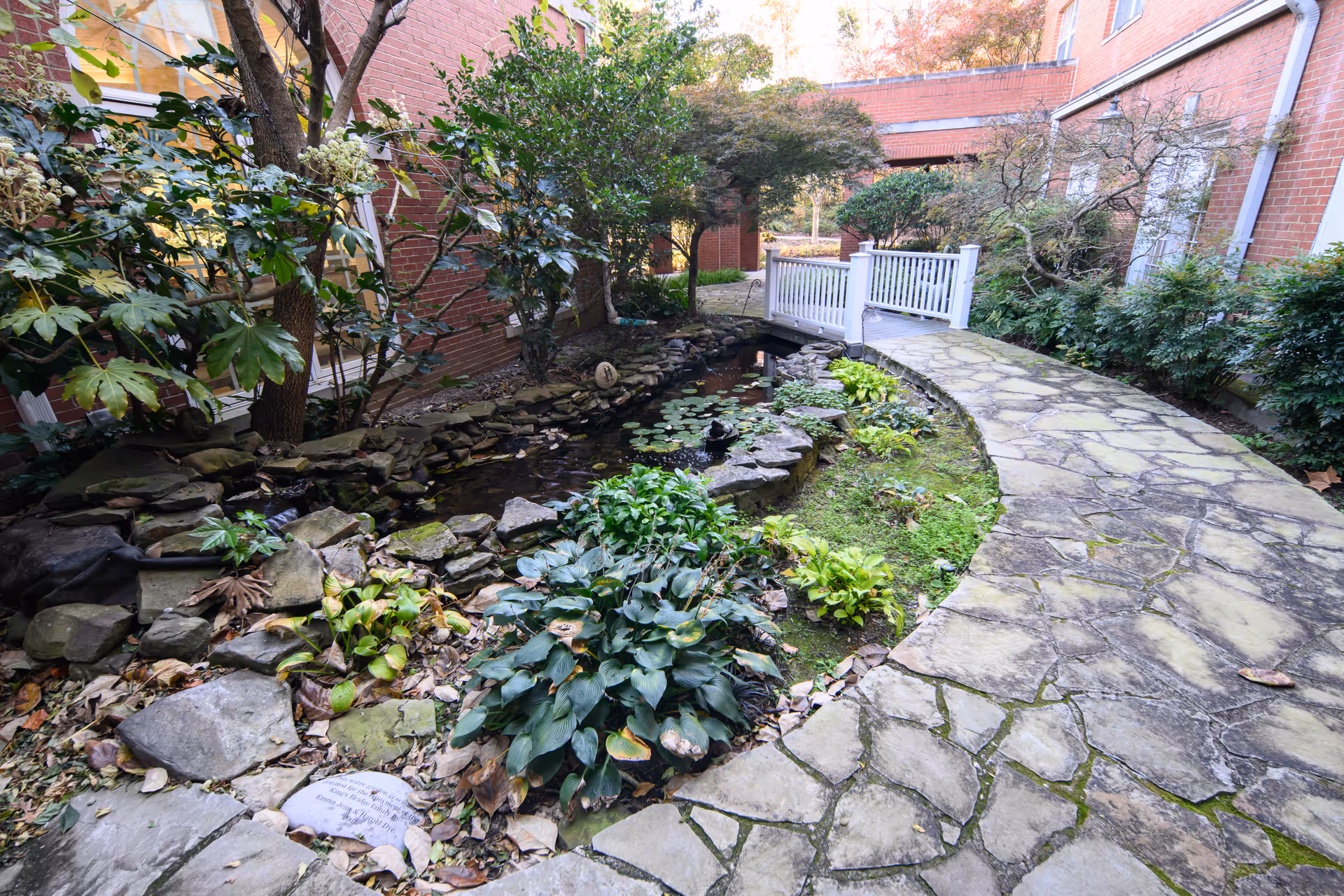 A landscaped courtyard with a curved stone walkway next to a small pond, plants and a white footbridge between brick buildings.