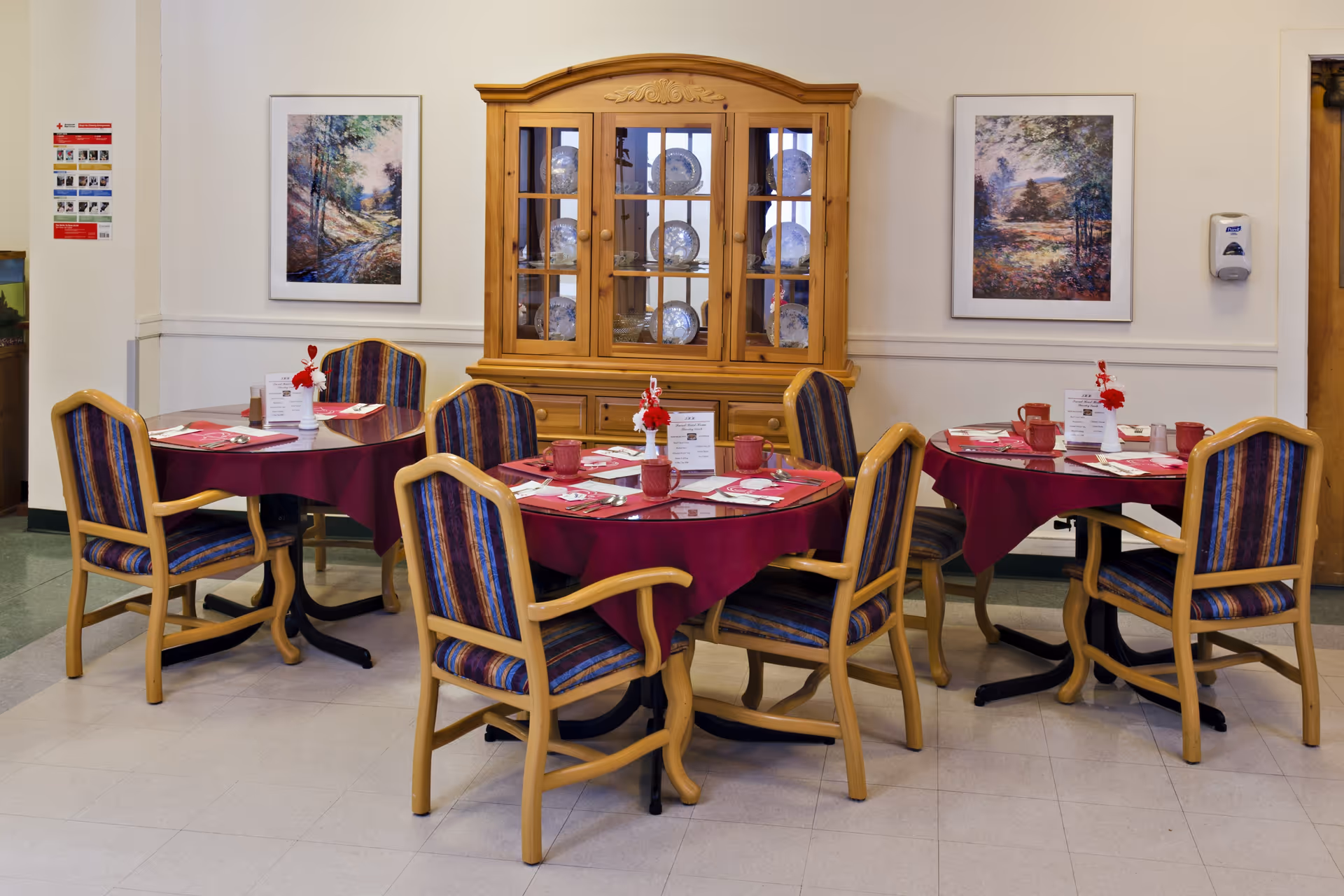A dining area with three round tables covered with burgundy tablecloths, each set with red mugs, napkins, and menus. The tables are surrounded by wooden chairs with blue and purple striped cushions. Behind the tables is a wooden cabinet with glass doors displaying decorative plates. Two framed landscape paintings hang on the wall, and a hand sanitizer dispenser is mounted on the right side.