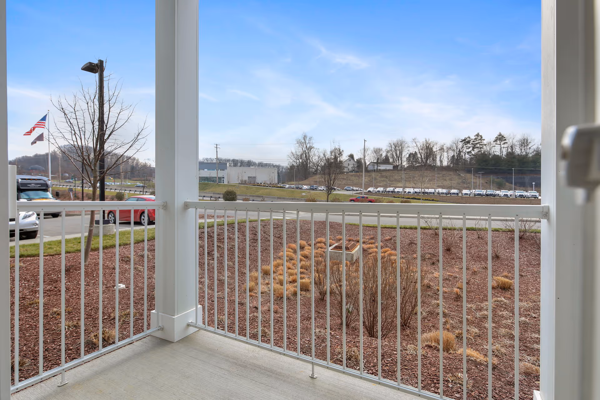 View from a small covered porch with white railing overlooking a landscaped mulch bed, a road with parked cars and flags under a blue sky.