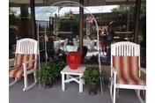 Two white rocking chairs with red and beige striped cushions flank a small white table holding a red decorative vase. Two potted plants are placed on the ground beside the table. The setting is on a covered patio or porch area with glass doors and windows reflecting the outdoor surroundings.