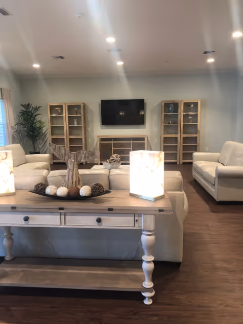 A well-lit living room with cream-colored sofas arranged around a wooden console table. The table holds decorative items including a wooden whale tail sculpture and spherical ornaments, along with two illuminated cube lamps. In the background, there are two wooden display cabinets flanking a wall-mounted flat-screen TV above a matching wooden media console. The room has wooden flooring and recessed ceiling lights.