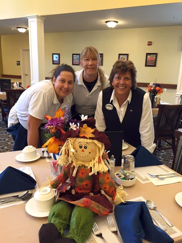 Three women smiling and posing behind a dining table set with plates, cups, utensils, and blue napkins. A colorful scarecrow doll and a floral arrangement are placed on the table. The setting appears to be a dining room in a senior living facility.