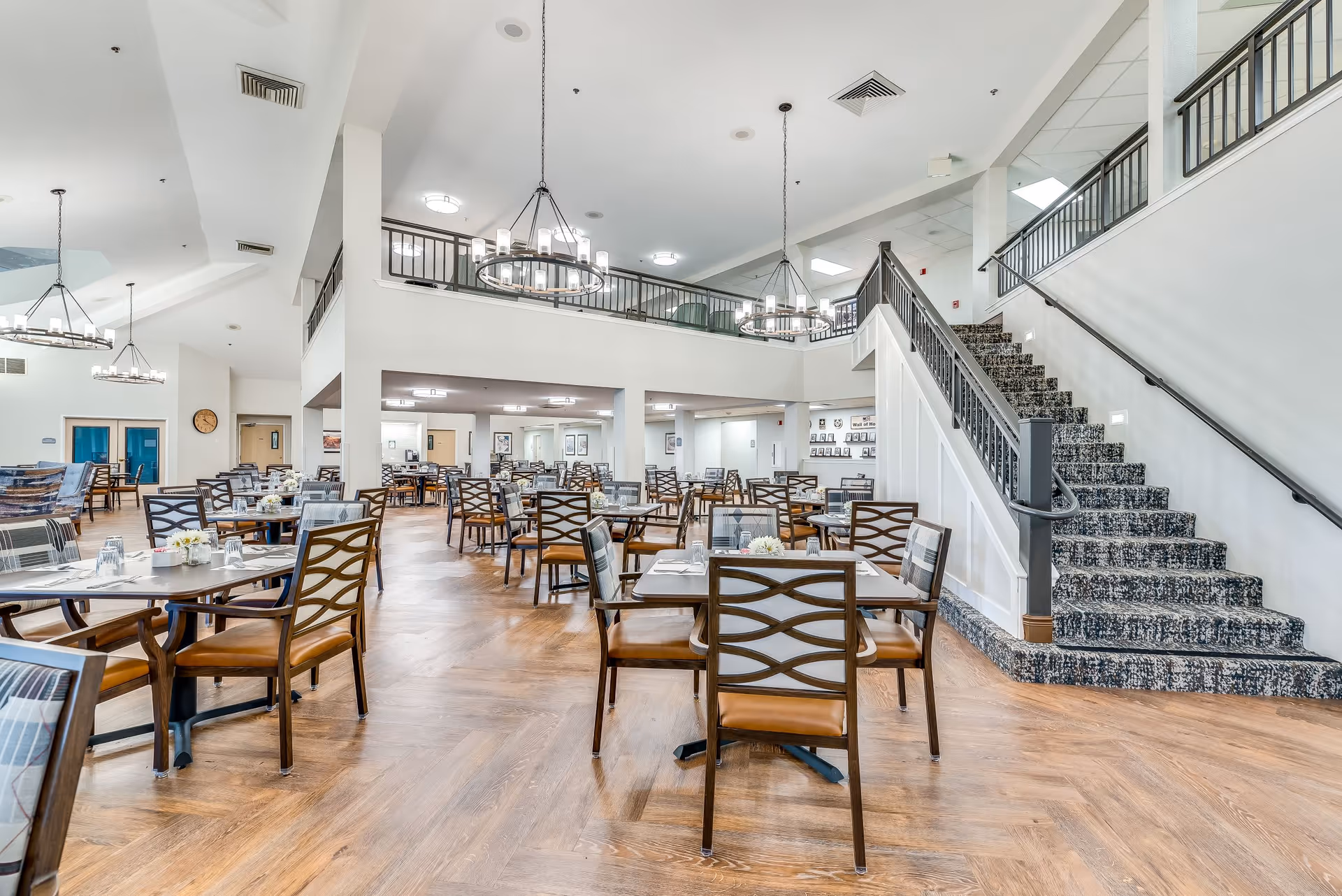 Spacious dining room in a senior living facility with multiple wooden tables and chairs arranged neatly. The room features high ceilings with modern chandeliers, a staircase with carpeted steps and black railing, and a mezzanine level overlooking the dining area. The floor is wooden with a herringbone pattern, and the walls are painted white, creating a bright and welcoming atmosphere.