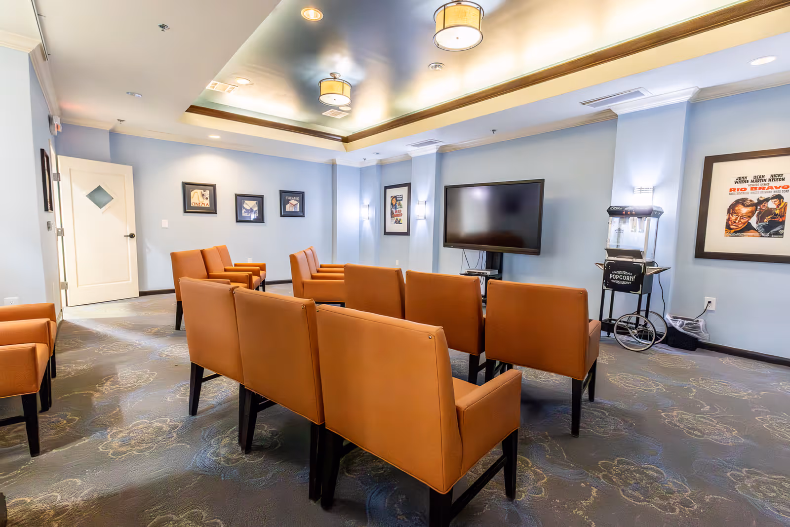 A cozy media room with rows of brown cushioned chairs facing a large flat-screen TV mounted on a light blue wall. The room has a popcorn machine on a stand to the right, framed movie posters on the walls, and soft ceiling lighting with recessed lights and hanging fixtures. The carpet has a floral pattern, and there is a white door on the left side of the room.