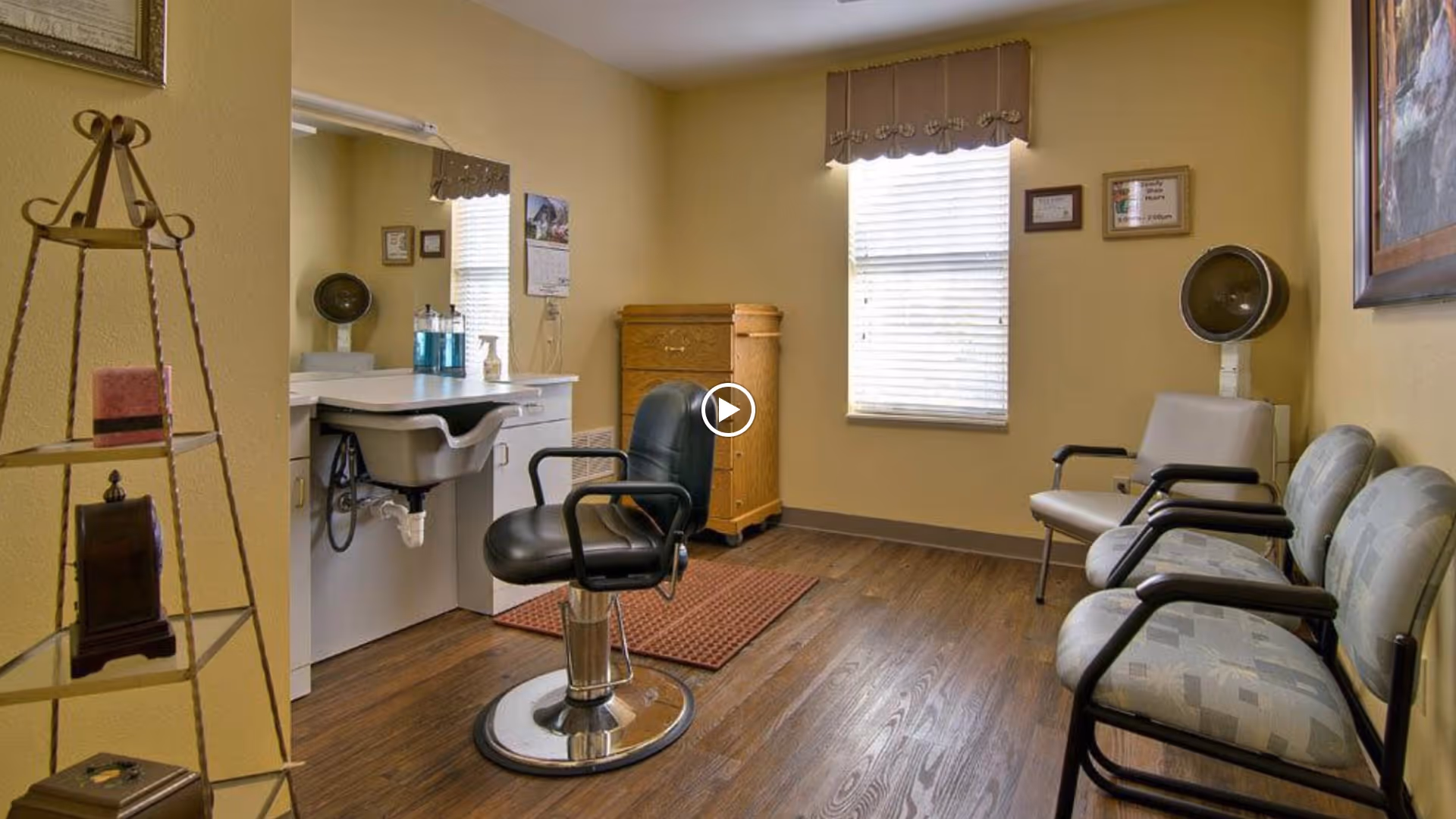 A small salon room with a black salon chair in front of a white sink and counter. There are three waiting chairs along the right wall, a hair dryer hood, a wooden cabinet, and a window with blinds and a valance. The floor is wooden, and the walls are painted yellow.