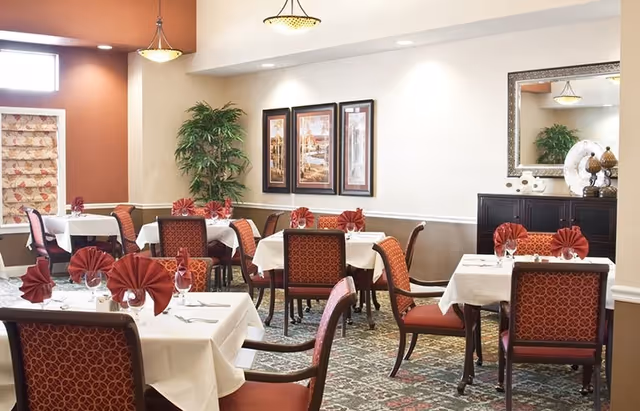 Bright, neatly arranged dining room with multiple tables set with white tablecloths, red fan-folded napkins, upholstered chairs, and wall art.