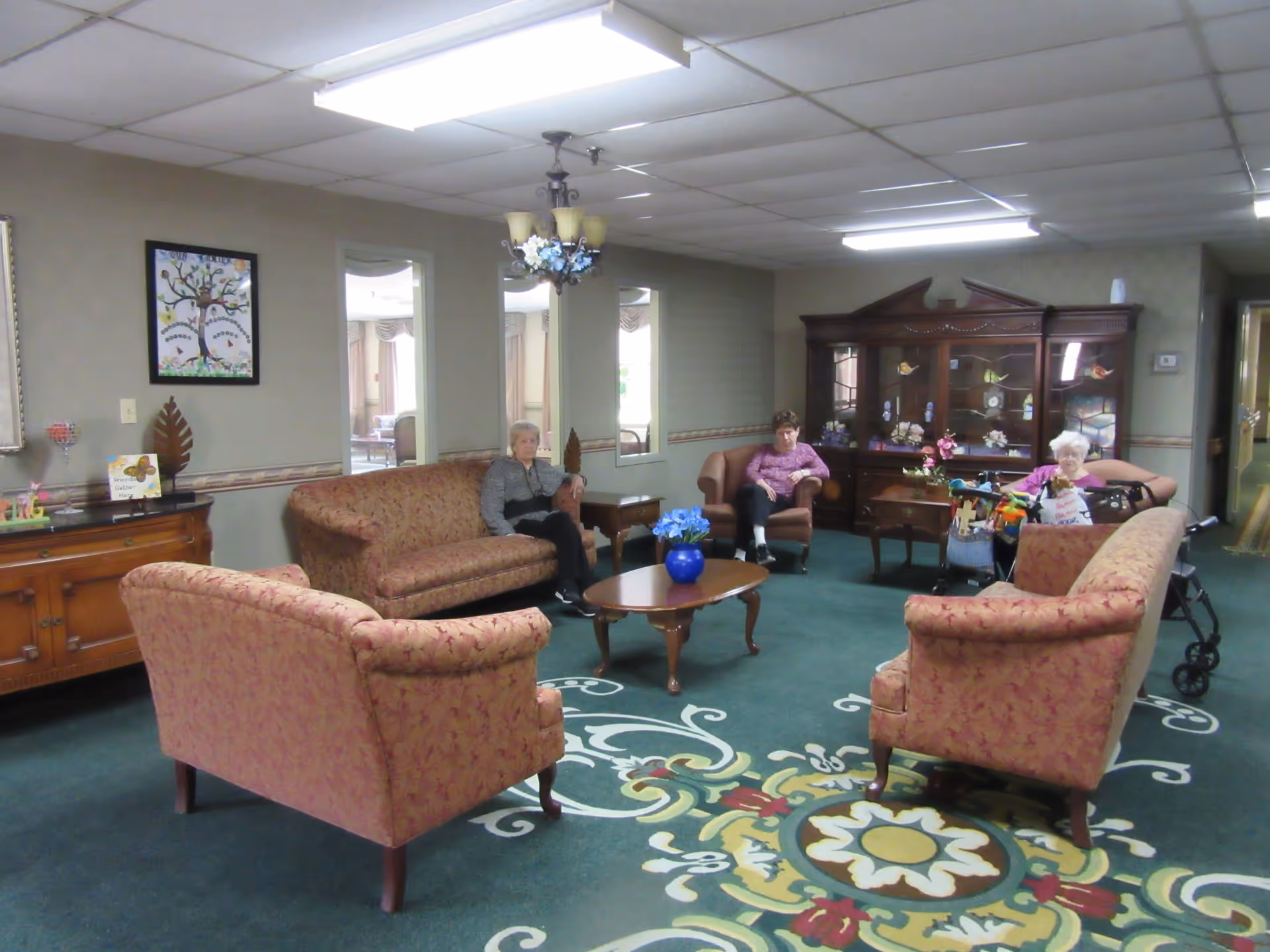 A common area in a senior living facility with three elderly women seated on upholstered chairs and sofas arranged around a wooden coffee table with a blue vase of flowers. The room has green carpet with a decorative pattern, beige walls, a wooden cabinet with glass doors displaying decorative items, and a chandelier hanging from the ceiling.